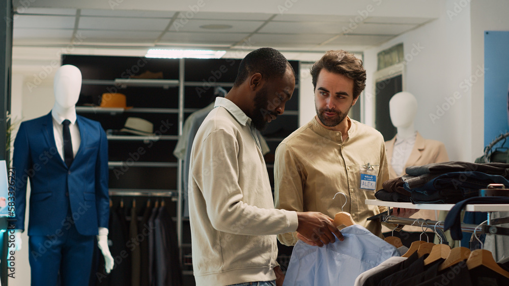 Fototapeta premium Young shopper asking shop worker for help with clothes, looking to buy formal wear from fashion boutique. Retail store assistant talking to customer about shopping for modern clothes.