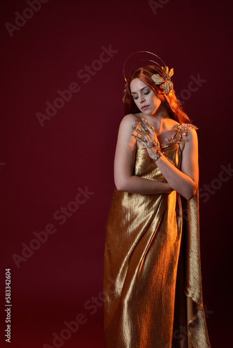 Close up fantasy portrait of beautiful woman model with red hair, goddess silk robes & gold crown. Posing with gestural hands reaching out, isolated on dark red studio background 