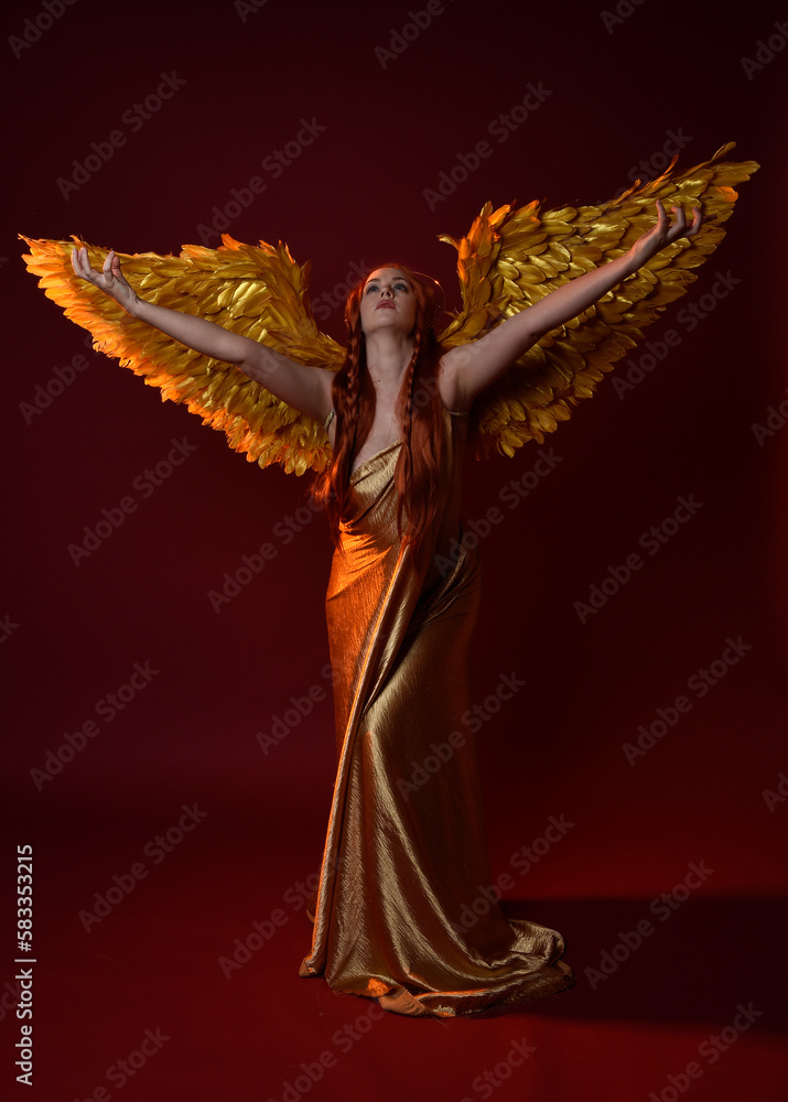 Full length portrait of beautiful woman model with long red hair, gold silk robes, crown & fantasy feather angel wings. Standing pose gestural hands reaching out isolated on dark red studio background