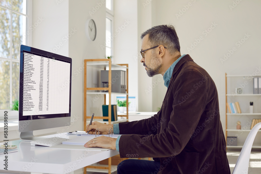 Portrait of serious, attentive, mature businessman man with glasses, sitting at his workplace and looking at his computer screen. Confident man taking notes in large journal for records.
