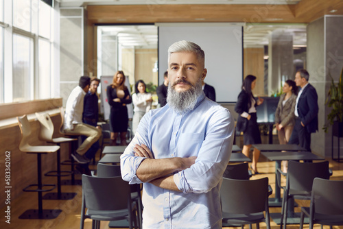Portrait of experienced team coach and business professional. Confident man with white hair and gray beard standing with arms folded in office after corporate training meeting with group of employees