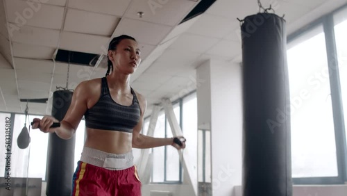 Young asian female boxer with braided hair fast and focused jumping rope next to punching bag warming up before boxing training in the indoor gym
