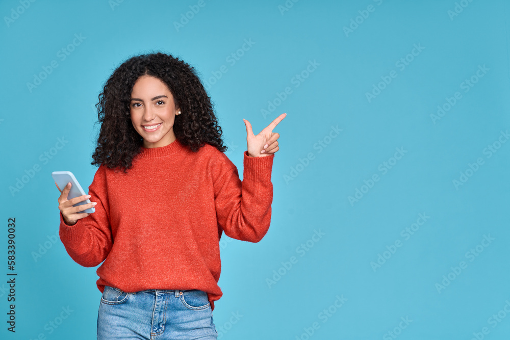 Young happy latin woman holding mobile phone pointing aside isolated on blue background. Smiling female model holding cellphone using cell presenting advertising new trendy shopping sale in app. Young happy latin woman holding mobile phone pointing aside isolated on blue background. Smiling female model holding cellphone using cell presenting advertising new trendy shopping sale in app.