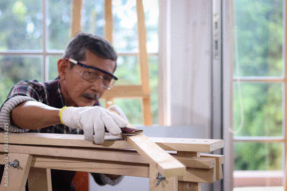 Senior Asian carpenter wear safety glasses and cloth gloves, sanding wood with sandpaper in woodwork carpentry, old citizen craftsman work in joinery workshop, elderly enjoy DIY hobby. Senior Asian carpenter wear safety glasses and cloth gloves, sanding wood with sandpaper in woodwork carpentry, old citizen craftsman work in joinery workshop, elderly enjoy DIY hobby.