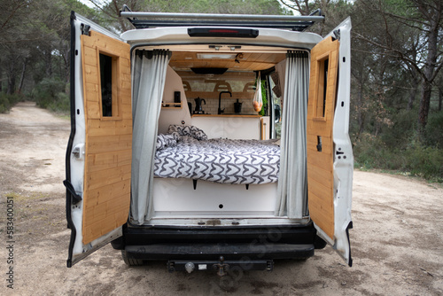 Back of camper with open doors, interior with wooden bed and kitchen, wooden doors, heart quilt and grey curtains, large white camper van, lonely forest interior.