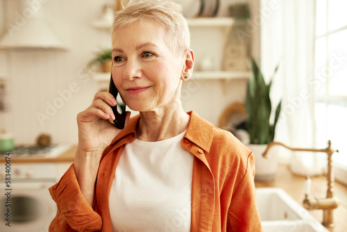 Happy mature lady with stylish short haircut having phone conversation standing at kitchen, talking to her beauty master, making an appointment for next week, listening attentively with smiling face