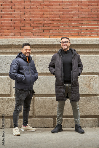 Two men in winter clothes looking at camera and smiling while standing outdoors on the street. Friendship concept.