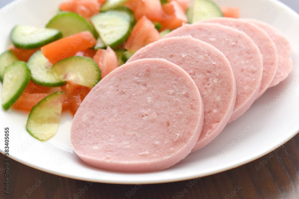 Greek Parizaki pork sausage slices with cucumber and tomato salad on a white plate. Very popular charcuterie products in Greece.  Sliced hams on a plate. 