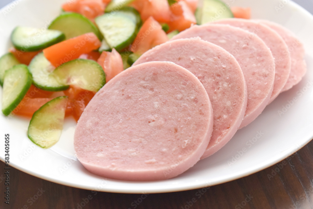 Greek Parizaki pork sausage slices with cucumber and tomato salad on a white plate. Very popular charcuterie products in Greece. Sliced hams on a plate. Greek Parizaki pork sausage slices with cucumber and tomato salad on a white plate. Very popular charcuterie products in Greece. Sliced hams on a plate.
