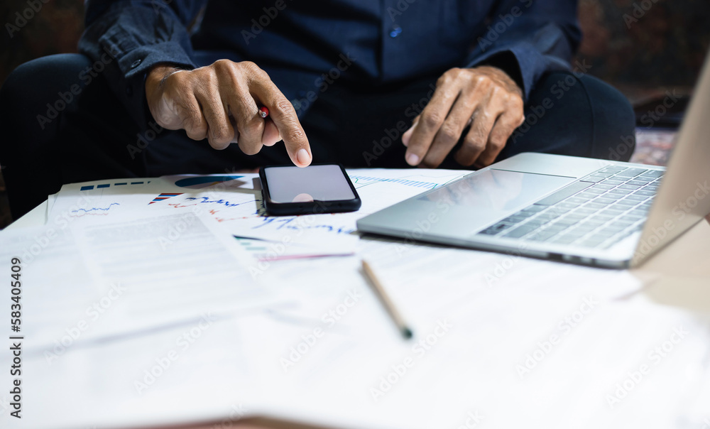 Asian businessman sitting on sofa at home using smartphone and laptop to check reports for investment plan via online meeting with business group, work from home concept Asian businessman sitting on sofa at home using smartphone and laptop to check reports for investment plan via online meeting with business group, work from home concept