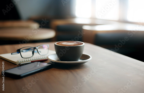 Close-up view, white cup of coffee with smartphone, notebook, pen and eye glasses on wooden table near bright window in cafe. Vintage light, blurred background