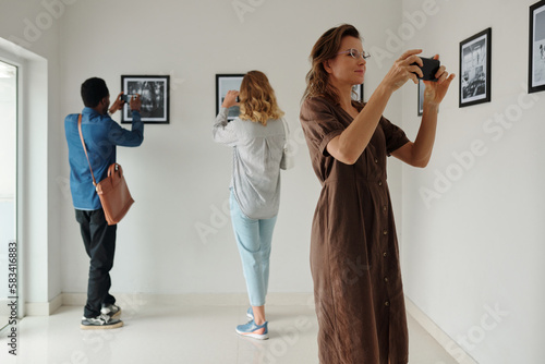 Woman in long brown cotton dress taking photo of artwork while standing in front of wall with pictures against young intercultural couple