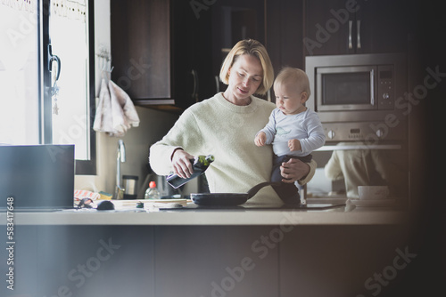 Happy mother and little infant baby boy together making pancakes for breakfast in domestic kitchen. Family, lifestyle, domestic life, food, healthy eating and people concept