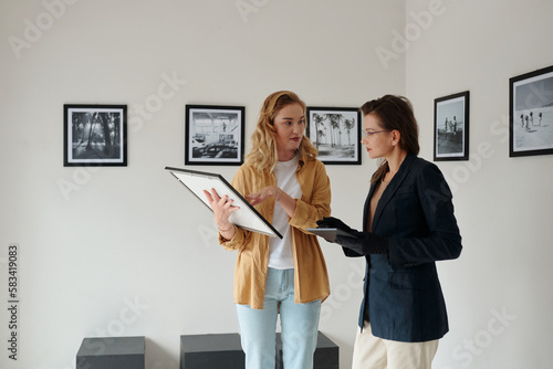 Two female workers of art gallery discussing restored picture and deciding where to hang it while preparing for opening of exhibition