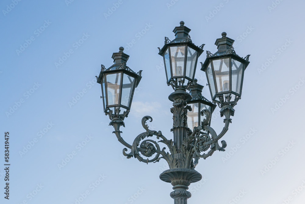 Old fashioned lamp. Vintage street lamp on a blue sky background. Street lamp on the background of the blue sky. Antique wrought iron lantern with glass inserts on the background of the blue sky.