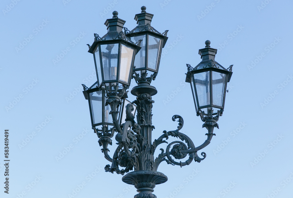 Old fashioned lamp. Vintage street lamp on a blue sky background. Street lamp on the background of the blue sky. Antique wrought iron lantern with glass inserts on the background of the blue sky.