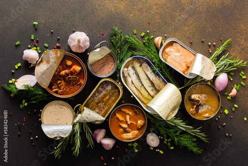 Foto Different open tin cans with canned fish among spices and herbs on a brown backg