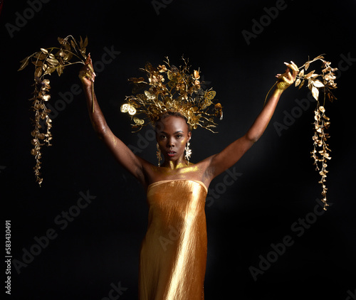  fantasy portrait of beautiful african woman model with afro, goddess silk robes and ornate floral wreath crown. gestural Posing holding golden flowers. isolated on dark studio background 