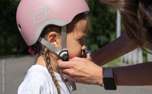 Mom putting on inline skates rollers protection helmet to daughter child in public park in summer. Family leisure outdoor sport activity game.