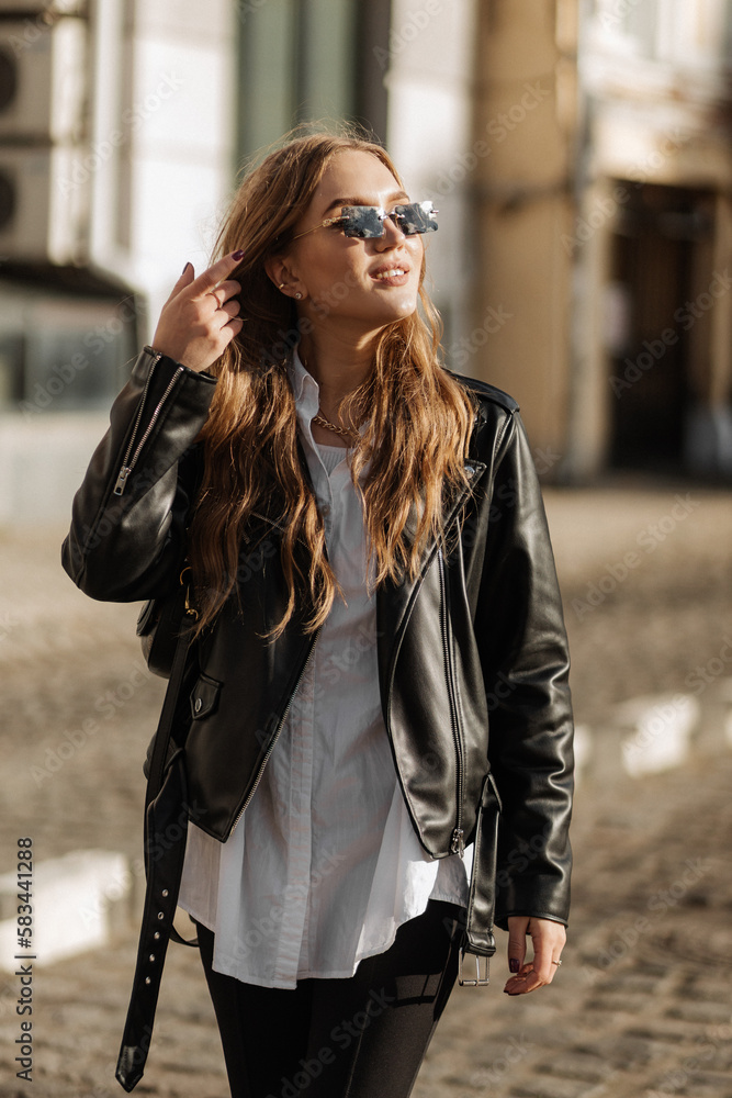 Fashionable  blonde woman model with  black leather jacket and style sunglasses walking the city street