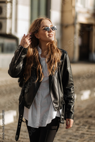 Fashionable blonde woman model with black leather jacket and style sunglasses walking the city street