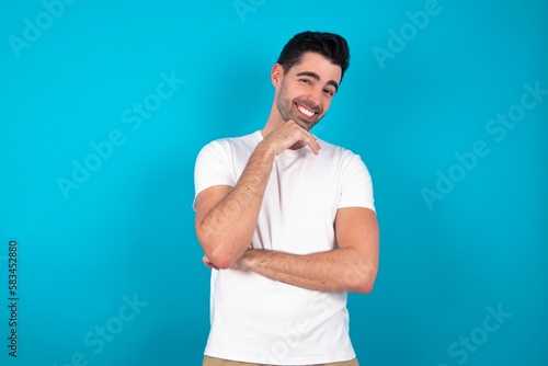 Optimistic Young man wearing white T-shirt over blue studio background keeps hands partly crossed and hand under chin, looks at camera with pleasure.