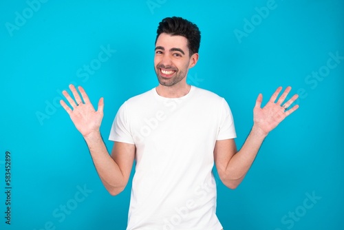 Optimistic Young man wearing white T-shirt over blue studio background raises palms from joy  happy to receive awesome present from someone  shouts loudly 