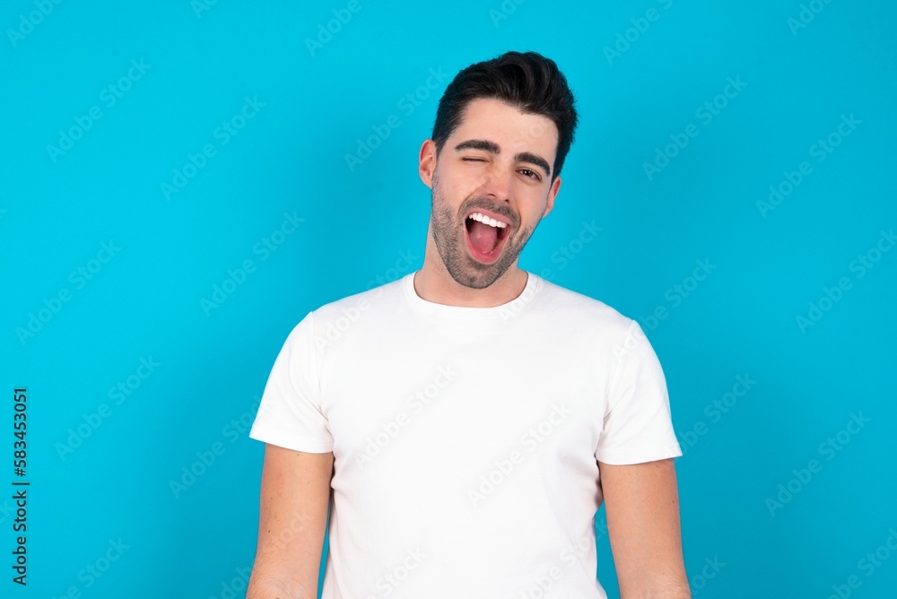 Young man wearing white T-shirt over blue studio background winking looking at the camera with sexy expression, cheerful and happy face. Young man wearing white T-shirt over blue studio background winking looking at the camera with sexy expression, cheerful and happy face.