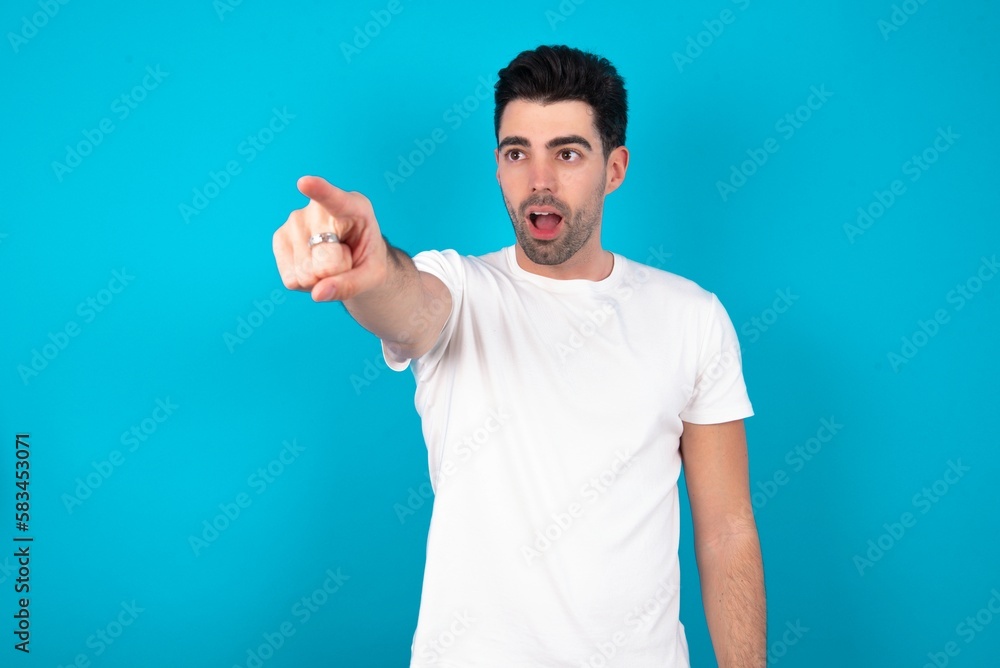 Young man wearing white T-shirt over blue studio background Pointing with finger surprised ahead, open mouth amazed expression, something on the front. Young man wearing white T-shirt over blue studio background Pointing with finger surprised ahead, open mouth amazed expression, something on the front.