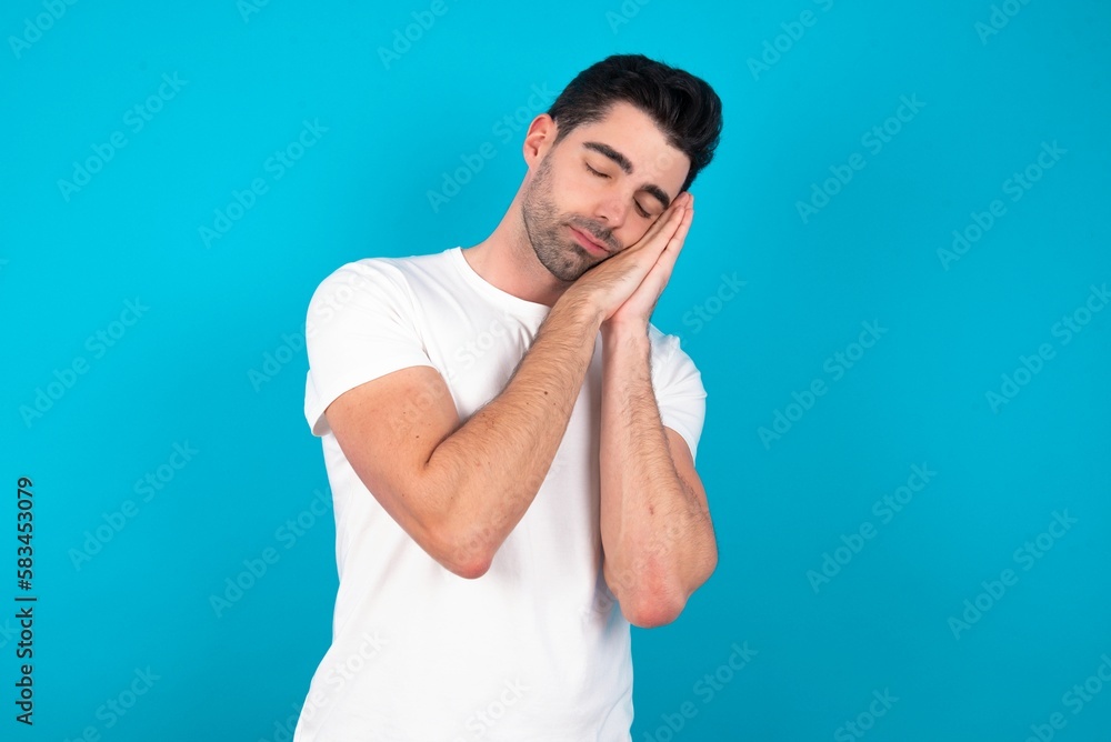 Young man wearing white T-shirt over blue studio background sleeping tired dreaming and posing with hands together while smiling with closed eyes. Young man wearing white T-shirt over blue studio background sleeping tired dreaming and posing with hands together while smiling with closed eyes.
