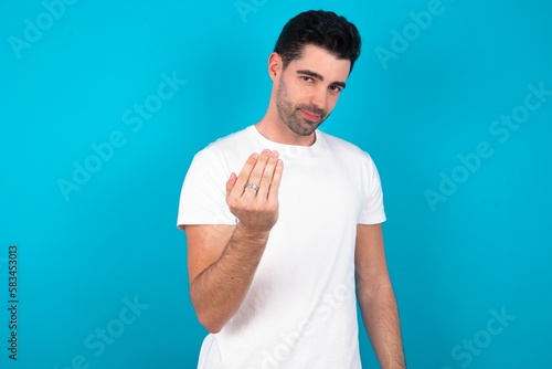 Young man wearing white T-shirt over blue studio background inviting to come with hand. Happy that you came