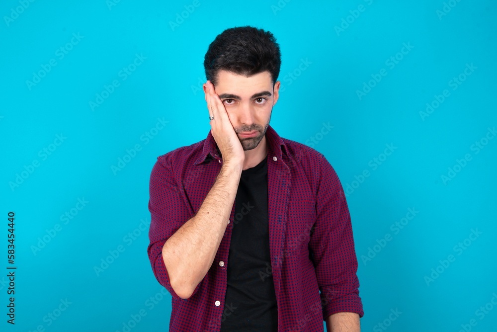 Sad lonely Young man standing over blue studio background touches cheek with hand bites lower lip and gazes with displeasure. Bad emotions Sad lonely Young man standing over blue studio background touches cheek with hand bites lower lip and gazes with displeasure. Bad emotions