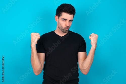 Irritated Young man standing over blue studio background blows cheeks with anger and raises clenched fists expresses rage and aggressive emotions. Furious model