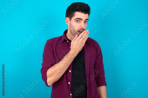 Young man standing over blue studio background covers mouth and looks with wonder at camera, cannot believe unexpected rumors.