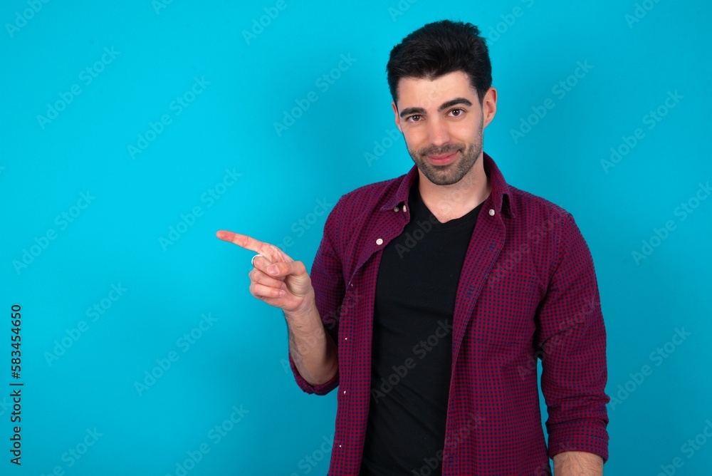 Young man standing over blue studio background points to side on blank space demonstrates advertisement. People and promotion concept Young man standing over blue studio background points to side on blank space demonstrates advertisement. People and promotion concept