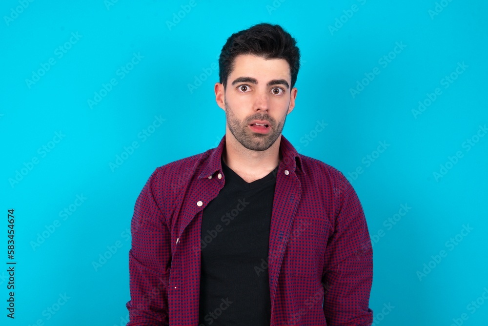 Obraz premium Shocked Young man standing over blue studio background stares bugged eyes keeps mouth opened has surprised expression. Omg concept