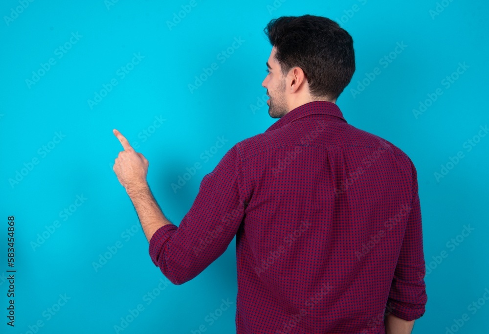 Young man standing over blue studio background pointing to object on copy space, rear view. Turn your back Young man standing over blue studio background pointing to object on copy space, rear view. Turn your back