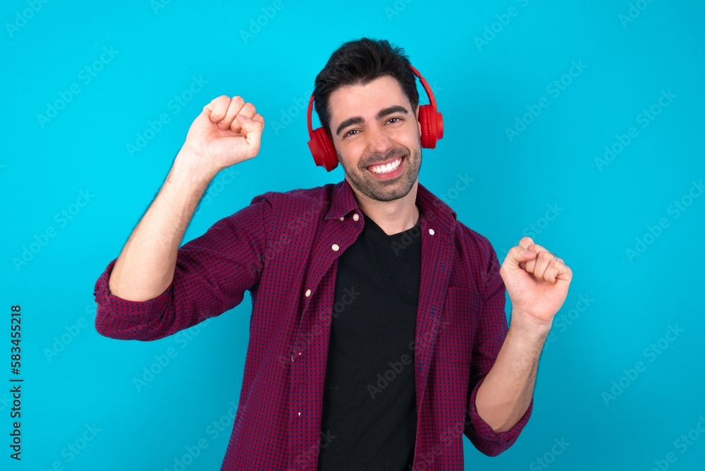 Carefree Young man standing over blue studio background with toothy smile raises arms dances carefree moves with rhythm of music listens music from playlist via headphones Carefree Young man standing over blue studio background with toothy smile raises arms dances carefree moves with rhythm of music listens music from playlist via headphones