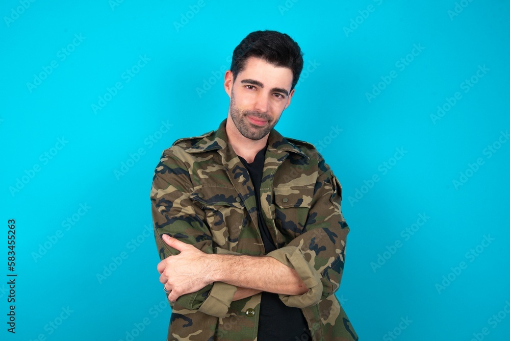 Portrait of charming Young man standing over blue studio background standing confidently smiling toothily with hands folded