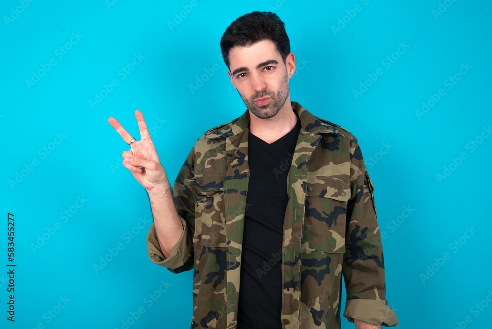 Young man standing over blue studio background makes peace gesture keeps lips folded shows v sign. Body language concept Young man standing over blue studio background makes peace gesture keeps lips folded shows v sign. Body language concept