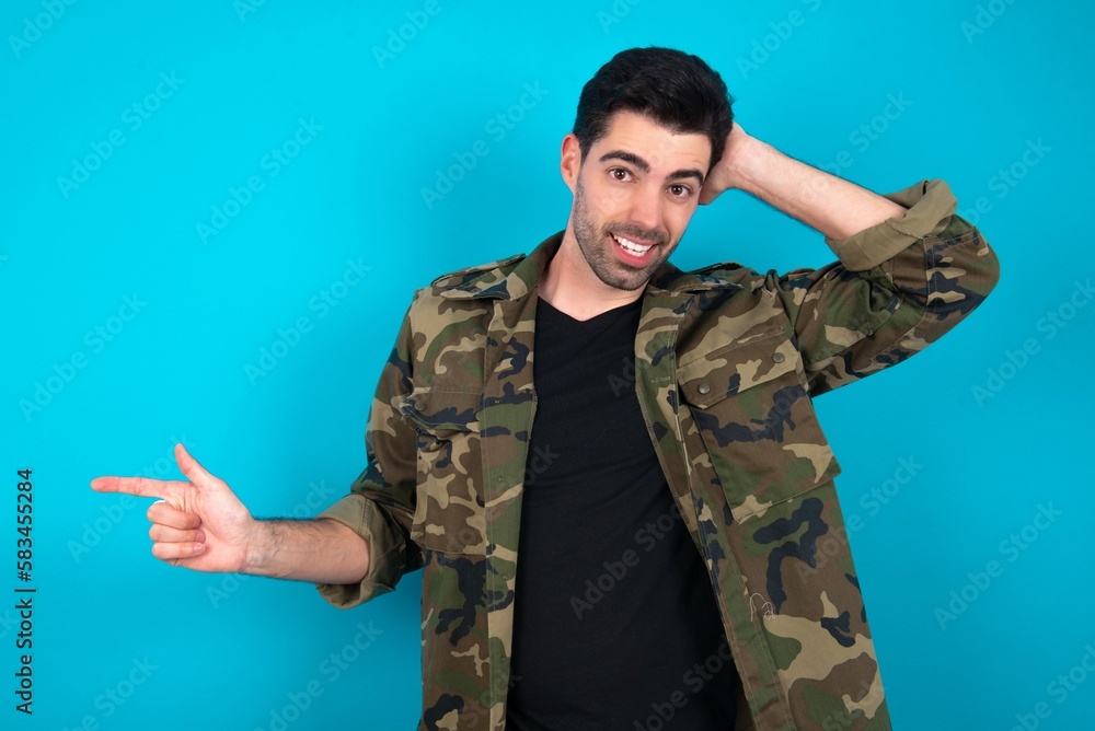 Surprised Young man standing over blue studio background pointing at empty space holding hand on head