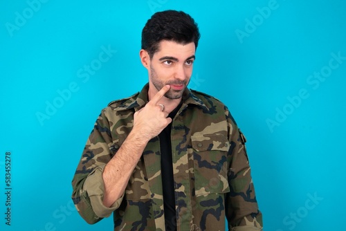 Lovely dreamy Young man standing over blue studio background keeps finger near lips looks aside copy space.