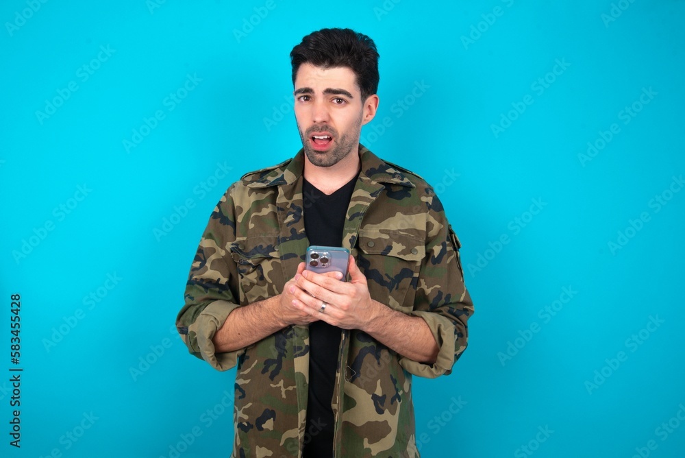 Photo of interested Young man standing over blue studio background hold telephone look side empty space screen