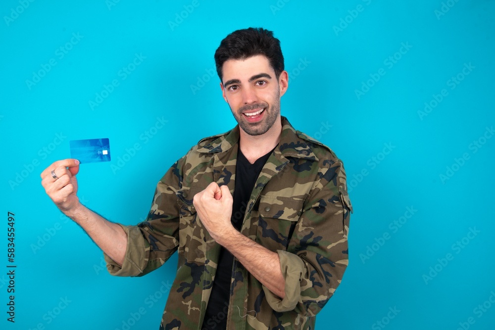 Excited happy positive cheerful smiling Young man standing over blue studio background hold credit card raise fist in victory