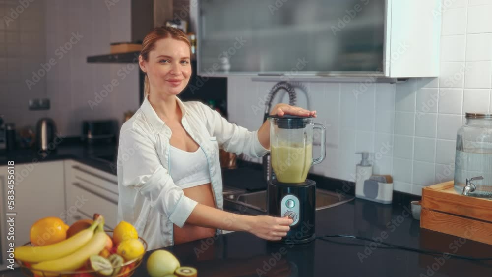 A young pregnant woman is preparing a smoothie with a blender in the kitchen. A pregnant woman