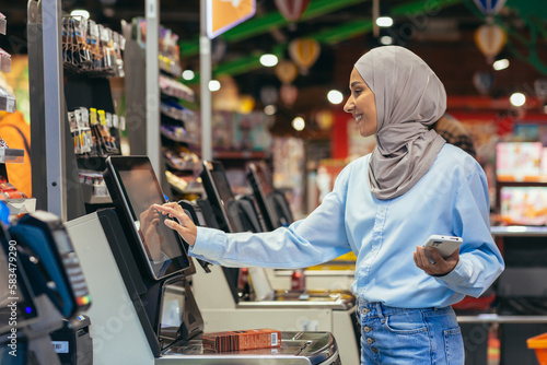A woman buyer in a supermarket in a hijab pays for goods at a self-service checkout, convenient service for customers.