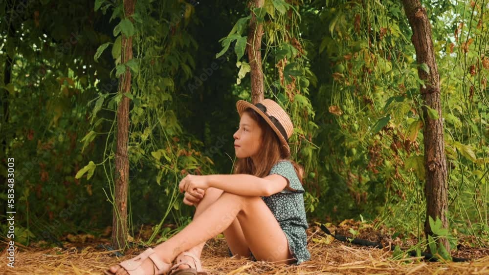 A cute smiling girl in a straw hat is sitting on the grass in the garden. Zoom in. Passion fruit plantations in the background. A cute smiling girl in a straw hat is sitting on the grass in the garden. Zoom in. Passion fruit plantations in the background.