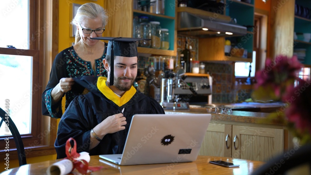 As part of a virtual commencement ceremony, a mother hooding her son graduating in front of laptop computer in their home dining room.