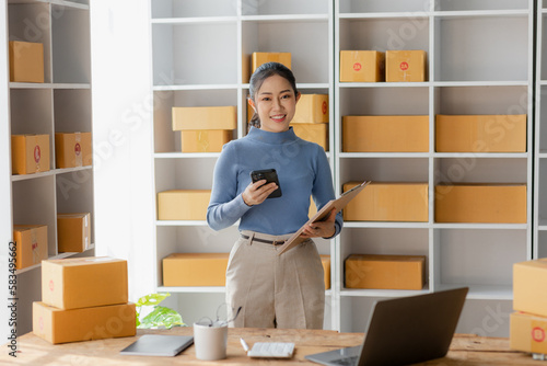 Starting an SME, an independent Asian female small business entrepreneur using a laptop and a smart phone. Cheerful success, cute smiley face. Asian woman raising her hand, packaging box, online marke