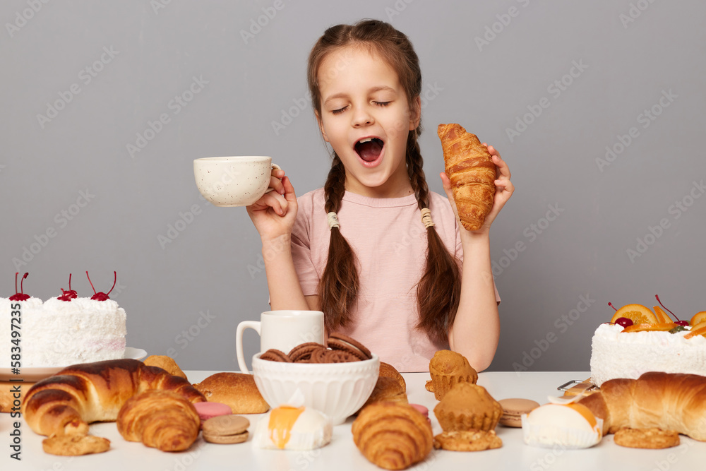 Portrait of satisfied overeating little girl with pigtails sitting at table isolated over gray background, holding cup with tea and croissant, feels sleepy, yawning. Portrait of satisfied overeating little girl with pigtails sitting at table isolated over gray background, holding cup with tea and croissant, feels sleepy, yawning.